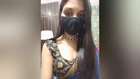 Video of a South Asian woman with long dark hair, wearing a black face mask, black and gold-patterned blouse, silver choker, and a green floral background.