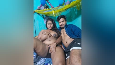 Video of a plus-size woman with large breasts, nude, and a slender man in a blue shirt and shorts, sitting on a bed with festive decorations in the background.