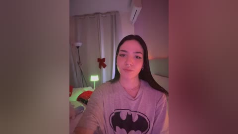 Media: Video of a young woman with long dark hair, wearing a Batman T-shirt, sitting on a bed with a red and green blanket, in a dimly lit bedroom with a fan and air conditioner.
