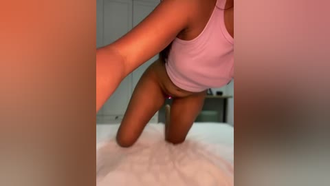 A video of a dark-skinned woman kneeling on a white bed, wearing a pink crop top and purple thong, in a dimly lit bedroom with white walls and a blurred background.