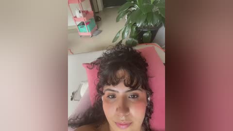 Video of a young woman with curly brown hair and light brown skin lying on a pink bed, surrounded by a green plant and pink furniture.