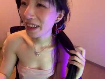 Media: A video of an East Asian woman with long black hair, wearing a strapless top, smiling, and holding her hair. The background is a plain beige wall.