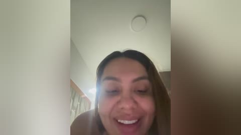 A close-up video of a smiling woman with medium-length brown hair and light brown skin, taken indoors, with a blurred background.