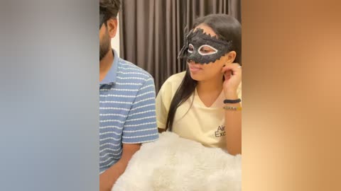 Video of a young South Asian couple, the woman with long dark hair, wearing a black Venetian mask, sits on a white fur blanket, while the man in a blue and white striped shirt stands beside her.