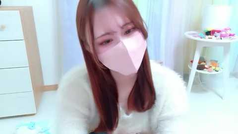 A video of an Asian woman with long brown hair, wearing a white mask and a fluffy white robe, seated indoors with a white dresser, colorful toys, and a lamp in the background.