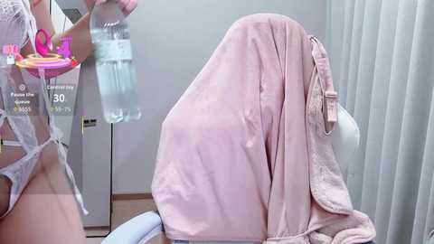 Video of a woman in a hospital gown holding a plastic cup of water, sitting on a bed with a pink blanket draped over her lap.