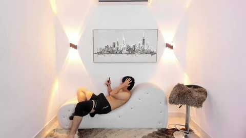 Media: Video of a topless Asian man in black shorts, lying on a white tufted sofa, playing a video game, surrounded by minimalist decor with modern lighting and cityscape artwork.