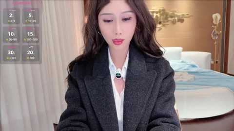 Video of a young East Asian woman with long, wavy brown hair, wearing a gray blazer over a white blouse, in a modern bedroom with a white bed, gold decor, and digital clock overlay.