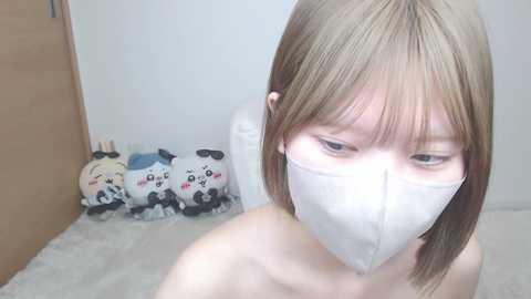 Media: Video of a young, slender Asian woman with short, light brown hair, wearing a white surgical mask and a white t-shirt. Background includes plush toys of pandas and a wooden door.