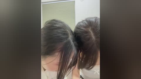 A video of two young Asian girls with long black hair in high ponytails, facing each other in a dimly lit room with white walls and a beige door.