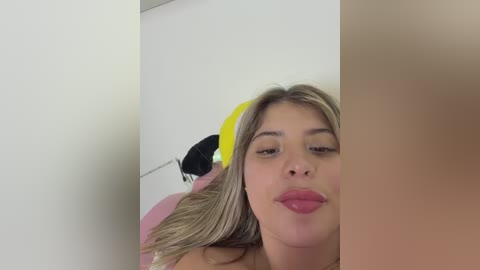 Video of a light-skinned woman with long blonde hair, wearing a yellow headband, blowing a kiss. Background includes a pink balloon and white wall.