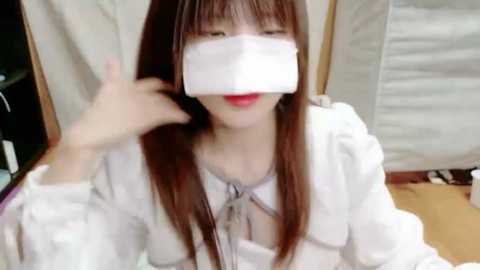 Video of a young Asian woman with long brown hair, wearing a white mask, white blouse, and sitting indoors with beige curtains and wooden floor.