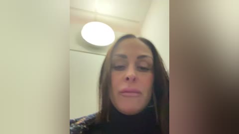 A video of a woman with light skin and long brown hair, wearing a black turtleneck, standing indoors. Background features a white wall, a ceiling light, and a blurred door.