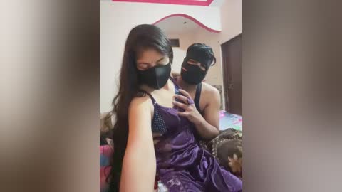 Video of a young woman with long dark hair in a purple saree, wearing a black mask, sitting on a bed with a man behind her, both smiling.
