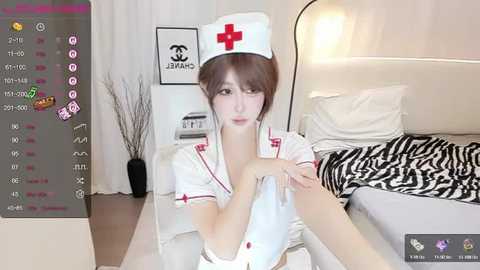 Media: Video of a young Asian woman in a nurse outfit, wearing a white cap with a red cross, in a modern bedroom with a bed, white curtains, and a zebra-patterned blanket.
