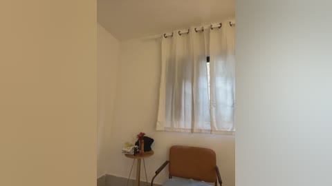 Media: Video of a sparsely furnished room with off-white walls, a single window with sheer white curtains, a wooden chair, and a small wooden table holding a red flower arrangement.