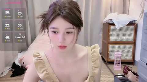 A video of an Asian woman with pale skin and dark hair tied back, wearing a beige dress. She sits at a table with a smartphone, a pink vibrator, and a thermometer. The background includes a wicker basket and a partially visible bed.