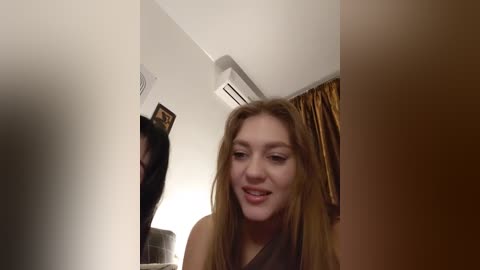 Video of a young woman with long, straight brown hair, wearing a dark top, smiling while looking at a mirror in a dimly lit room with cream walls, a brown curtain, and a ceiling air conditioner.