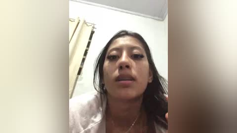 Media: Video of a young Asian woman with wet hair, wearing a light-colored robe, standing in a bathroom with a beige curtain. Her expression is neutral.