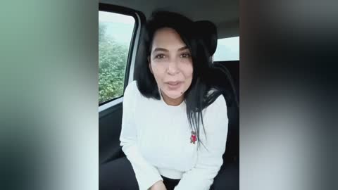 Video of a woman with long black hair, fair skin, and a white long-sleeved shirt, seated in a car with greenery outside the window.