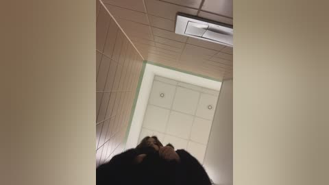 Video of a narrow, beige-tiled corridor with a green border. A person's head and shoulders are visible in the foreground, wearing a black jacket. The ceiling has rectangular fluorescent lights. The scene conveys a sense of confinement and minimalism.