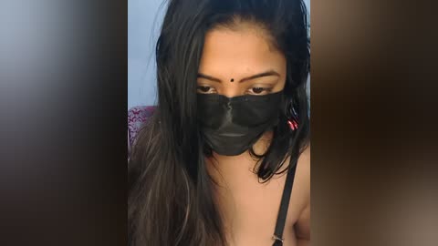 Media: Video of a South Asian woman with long black hair, wearing a black face mask, a red bindi, and a black lace bra. She has a fair complexion and is indoors, with blurred background.