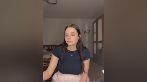 Video of a young Caucasian woman with long brown hair, fair skin, and a slim physique, wearing a dark blue shirt and pink skirt, standing in a modern, minimally decorated bedroom.