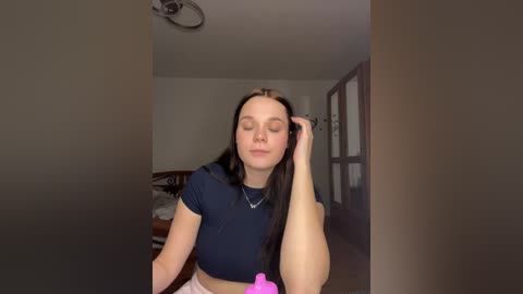 Media: A video of a young woman with long, straight black hair, fair skin, and a slender build, wearing a navy blue crop top, standing in a dimly lit bedroom with a bed and a door in the background.