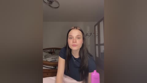 Media: Video of a young Caucasian woman with long, straight black hair, wearing a dark blue short-sleeved top, sitting on a bed in a dimly lit bedroom.