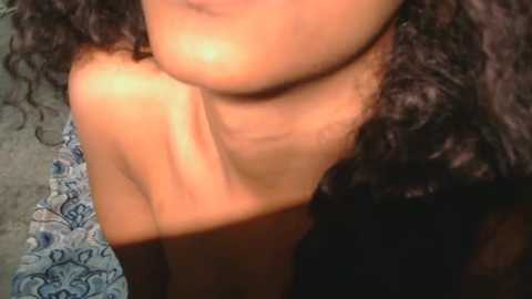 Video of a topless, light-skinned woman with curly dark hair, showcasing her bare breasts and collarbone. Background includes a patterned rug and a textured surface.