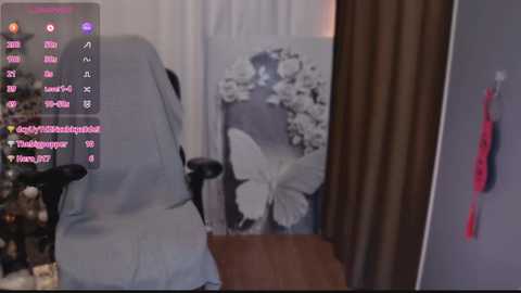 Video of a person in a gray chair with a black armrest, facing a white wall with a large black and white butterfly painting. Text on the left lists usernames and messages.
