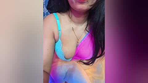 Video of a young Asian woman with long black hair, wearing a colorful tie-dye top and a turquoise lace bra, smiling slightly, indoors with a blurred background.