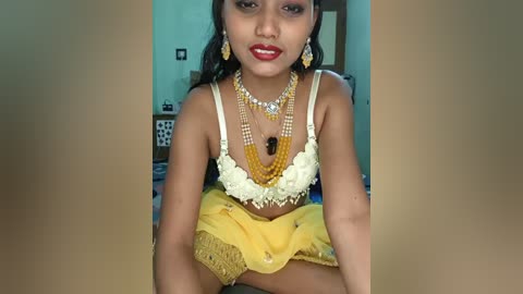 Video of a young South Asian woman with fair skin, dark hair, and red lipstick, wearing a yellow and white traditional outfit, adorned with gold jewelry, sitting in a simple indoor setting.