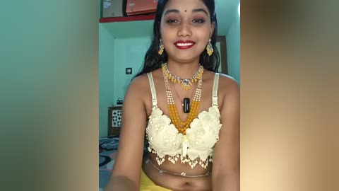 Video of a young woman with medium brown skin, wearing a yellow lace bra, multiple gold necklaces, and red lipstick, in a room with green walls and a wooden shelf.