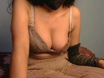 Media: Video of a woman with long black hair, wearing a gray lace bra and beige ribbed sweater. She has a black mask covering her face, and a black arm brace on her right arm, sitting on a bed with a patterned quilt.