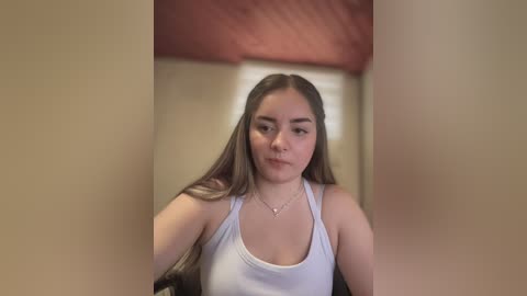 Media: Video of a young woman with long, straight brown hair, wearing a white tank top, standing in a dimly lit room with beige walls and a red ceiling.