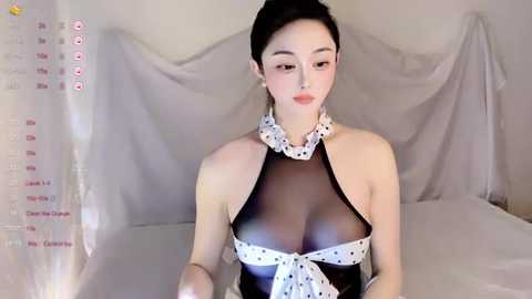 A video of an Asian woman with pale skin, dark hair, and a revealing black halter dress with white polka dots, standing in a bedroom with white sheets and drapes.