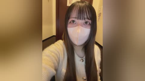 A video of an Asian woman with long, straight brown hair and bangs, wearing a white face mask, yellow cardigan, and white shirt, standing in a beige hallway with a wooden door in the background.