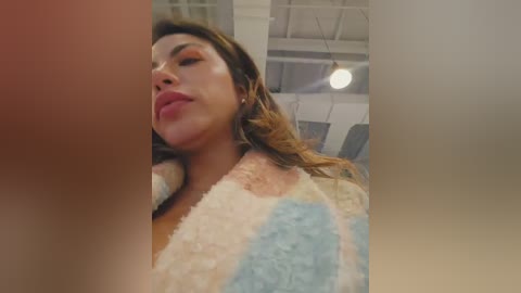 Video of a Latina woman with light brown skin, long brown hair, and full lips, wearing a pastel-colored fuzzy robe, captured in a modern, industrial-style room with white walls and exposed beams.