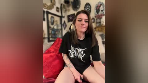 A video of a young woman with long brown hair, tattoos, and a black \"Support The Berserkers\" t-shirt, sitting on a red bedspread in a cluttered room with many decorative items.