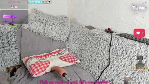 A video of a plush gray couch with a furry white throw, featuring a pink and white checkered pillow and a realistic-looking sex toy, set against a white brick wall.