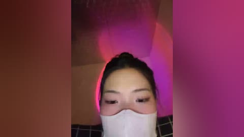 Video of an Asian woman with long black hair tied back, wearing a white surgical mask. She has makeup and is in a dimly lit room with pink and purple lighting.
