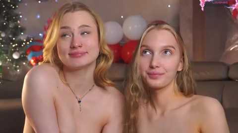 Video of two topless young women with fair skin, blonde hair, and playful expressions, seated on a couch; festive background includes Christmas decor and balloons.