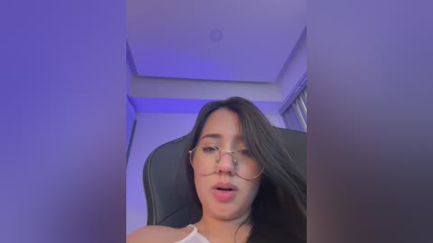 Video of a young woman with long black hair, wearing glasses and a white top, sitting on a black chair. The background is a modern room with purple lighting.
