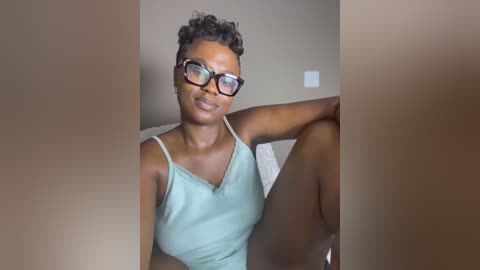 Video of a Black woman with short curly hair, wearing glasses and a light green tank top, sitting on a bed.