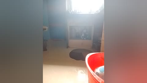 A video of a dimly lit, sparsely furnished room with a large, bright window, a blue plastic chair, and a red plastic bucket.