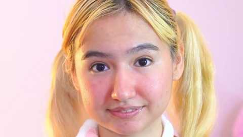 Media: Video of a young Asian woman with straight blonde hair in pigtails, wearing a white shirt, smiling softly against a pastel pink background.
