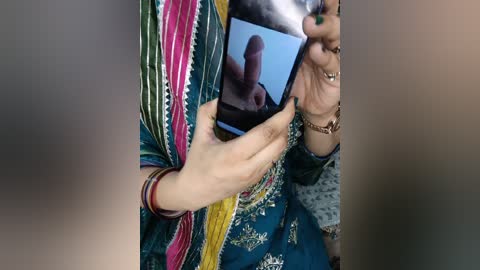 Media: A video captures a person holding a smartphone with a close-up image of a penis on the screen, wearing a colorful, patterned dupatta with gold embroidery, and a wristwatch.