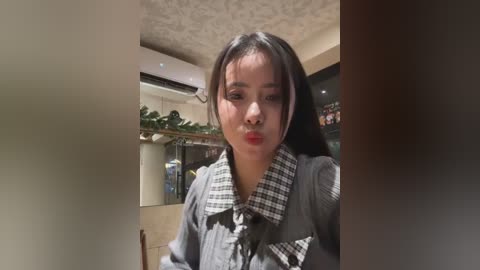 Video of a young Asian woman with long black hair, wearing a plaid shirt, puckering her lips in a pout, standing in a modern, warmly lit restaurant with beige walls and a ceiling fan.