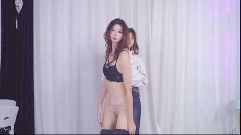 Video of two East Asian women in lingerie, one in black lace bra and panties, the other in a white blouse and jeans, standing against white and black curtains.